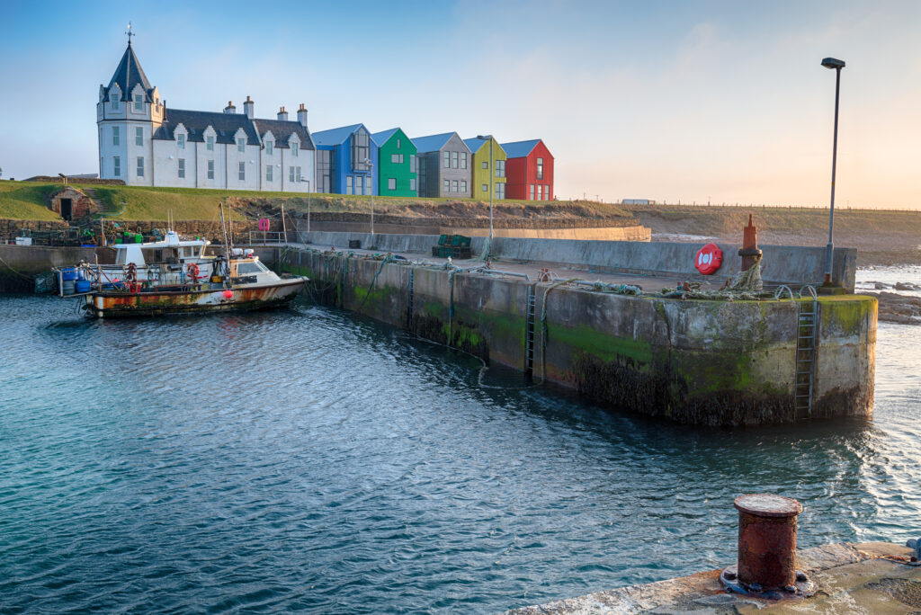 Fishing boat and colorful waterfront buildings at John o’ Groats Harbor in Scotland.