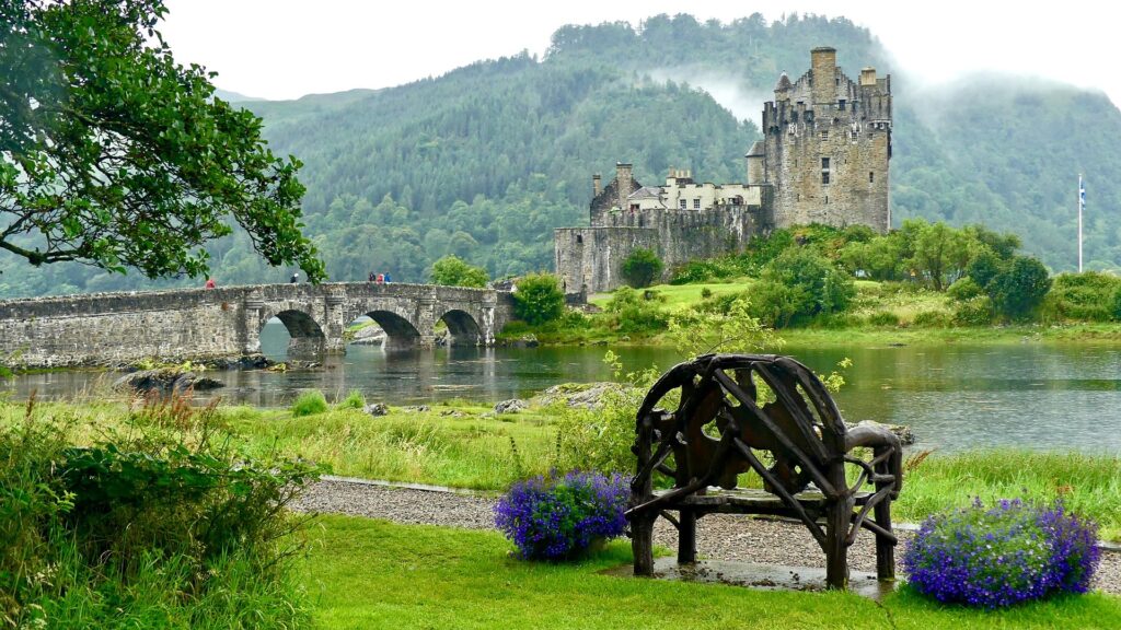 View of Eilean Donan Castle with bench flanked with purple flowers in the foreground.