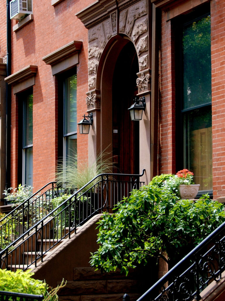 Brooklyn brownstone with an ornate arched entrance, front stoop, and flower-filled planters.